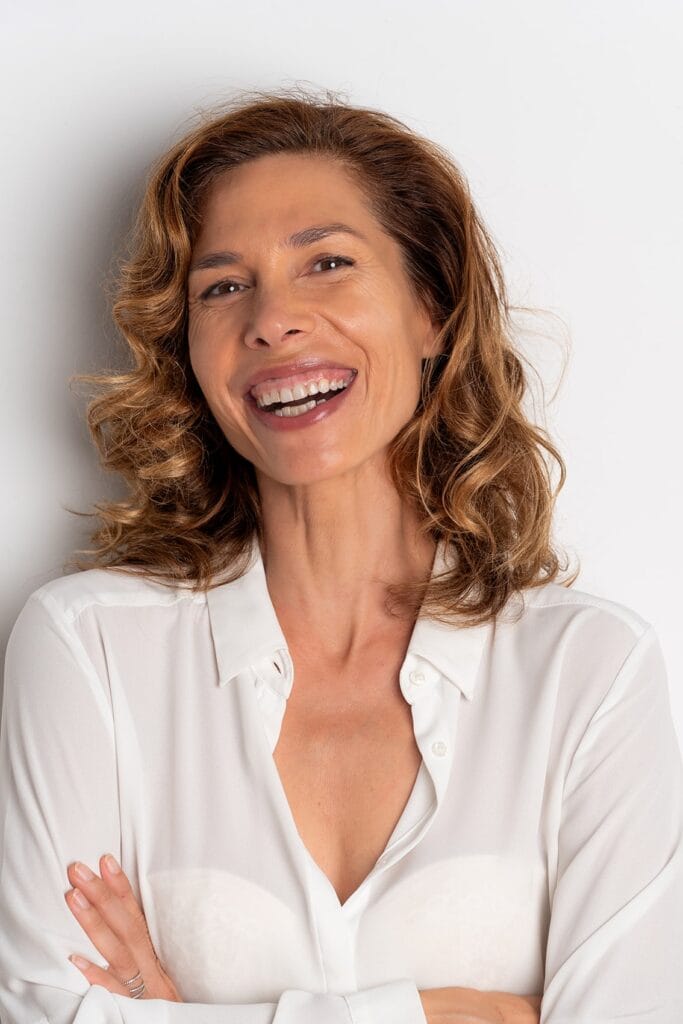 fotografo mallorca 2080estudio actores y actrices00001 fotografia-de-marca-personal-mallorca Mujer de pelo rizado con blusa blanca y brazos cruzados riendo sobre fondo claro: retrato profesional de marca personal en estudio en Palma