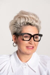 Primer plano de mujer con gafas de pasta negra, pelo corto rubio y blusa blanca