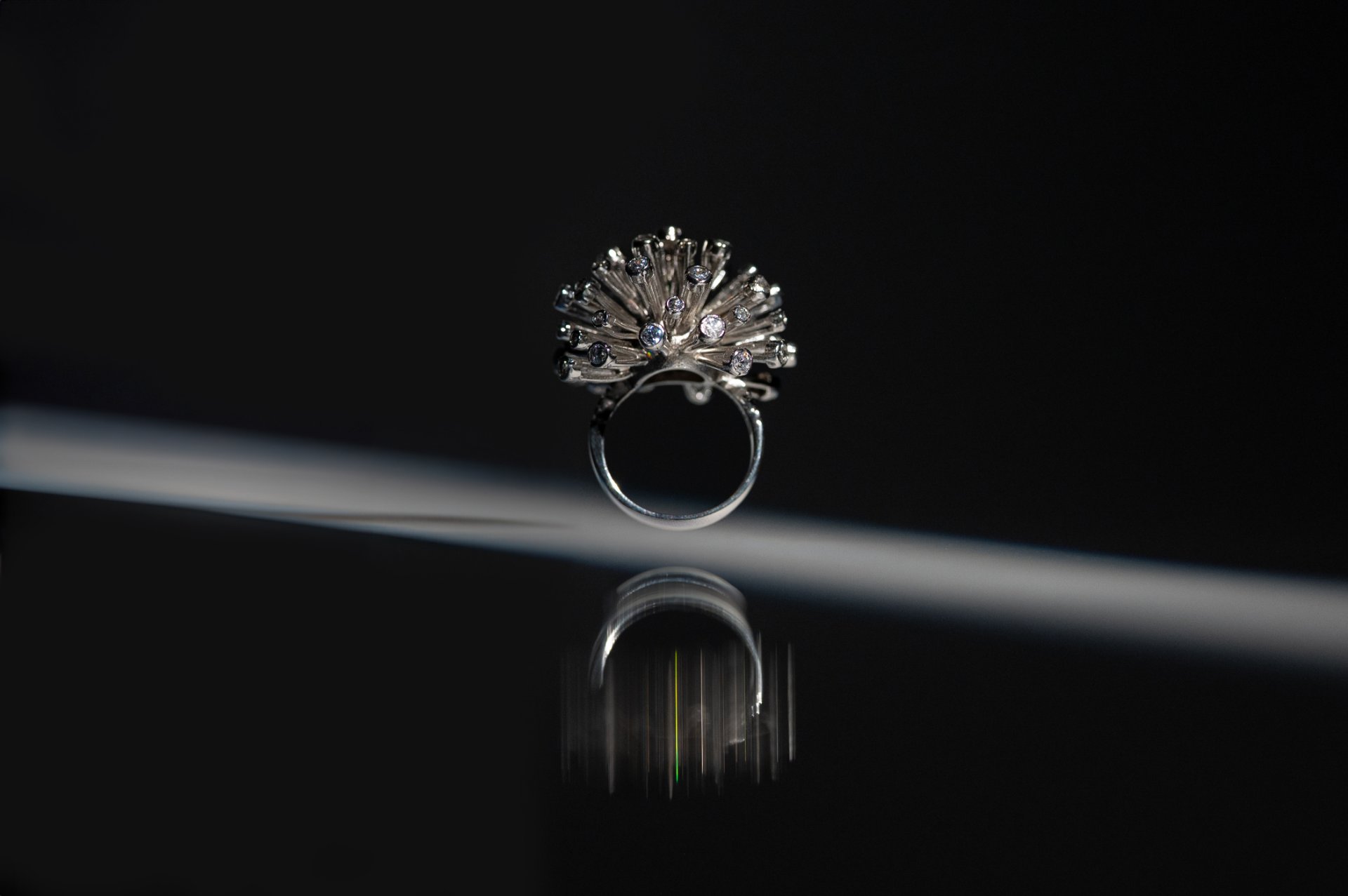 Anillo de plata con piedras sobre fondo negro, atravesado por un haz de luz y con su reflejo: fotografía de producto de joyería en Palma de Mallorca, por Flor Rajo